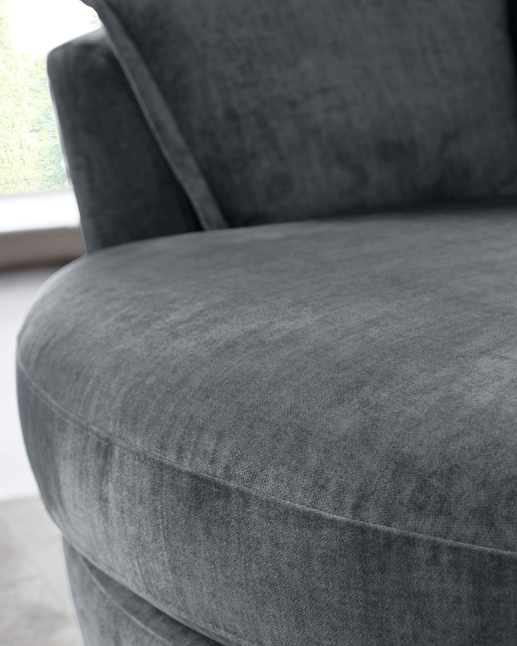 Close-up of a contemporary dark grey upholstered armchair with a plush seat cushion and soft fabric texture, partially visible armrest, with a blurred natural light background indicating a window.