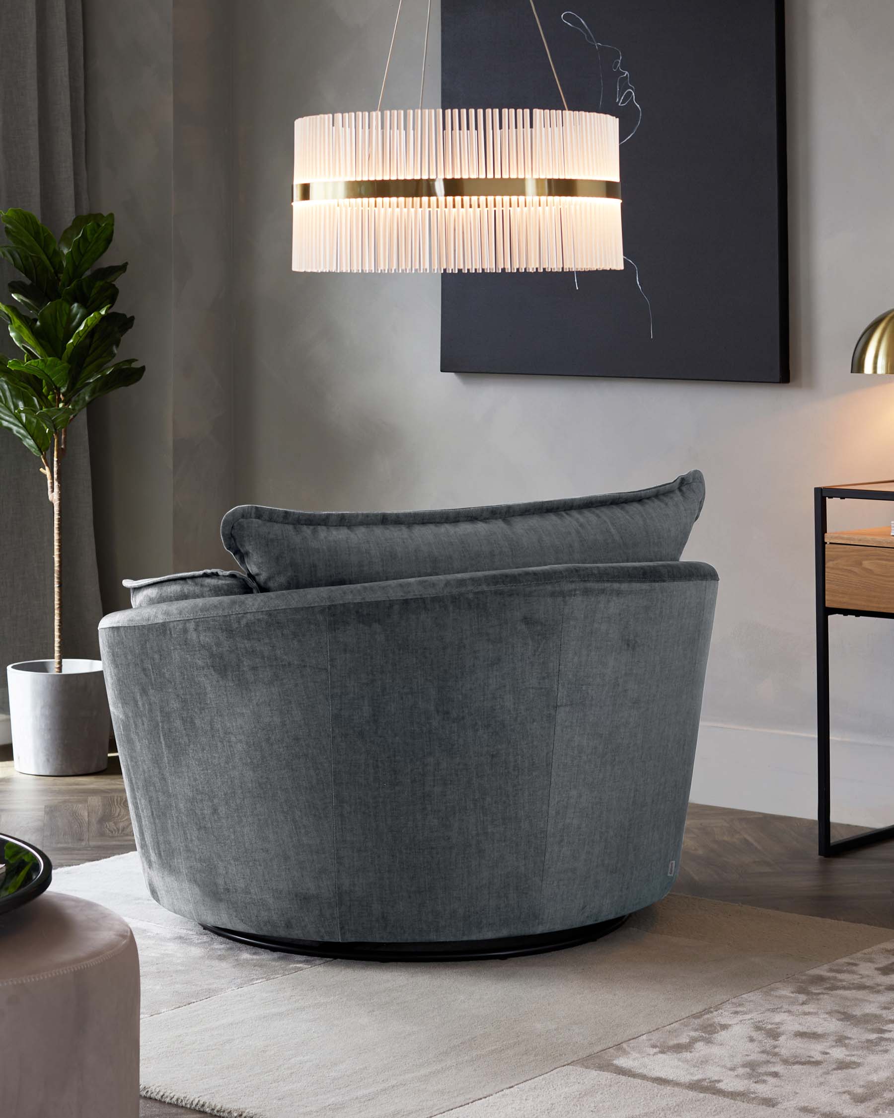 Plush circular velvet lounge chair in a muted grey tone with deep cushioning and a rounded back, resting on a low-profile black base. Adjacent to the chair is a minimalistic wooden side table with a simple, modern design and a small potted plant. Over the chair hangs a contemporary pendant light with vertical slats and ambient illumination, all set against a backdrop of a grey wall with subtle wall art and drapery.