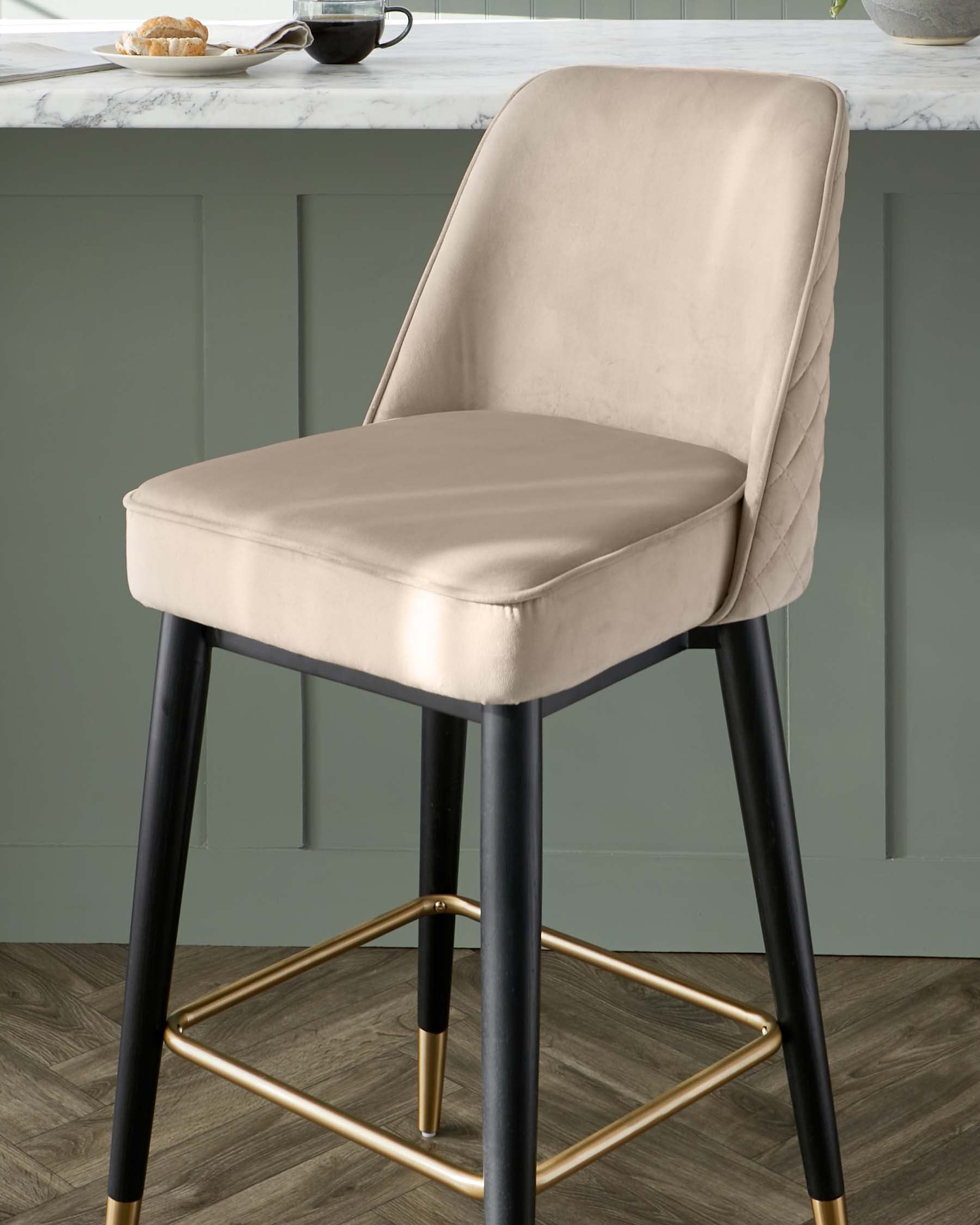 Elegant modern barstool with a taupe velvet seat and backrest featuring quilted detailing. The chair is supported by sleek black legs with gold footrest accents, set against a kitchen counter backdrop.