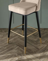 Elegant modern bar stool with black tapered legs accented with gold tips and a matching gold footrest. The seat is upholstered in a plush light beige fabric with diamond tufting on the backrest.