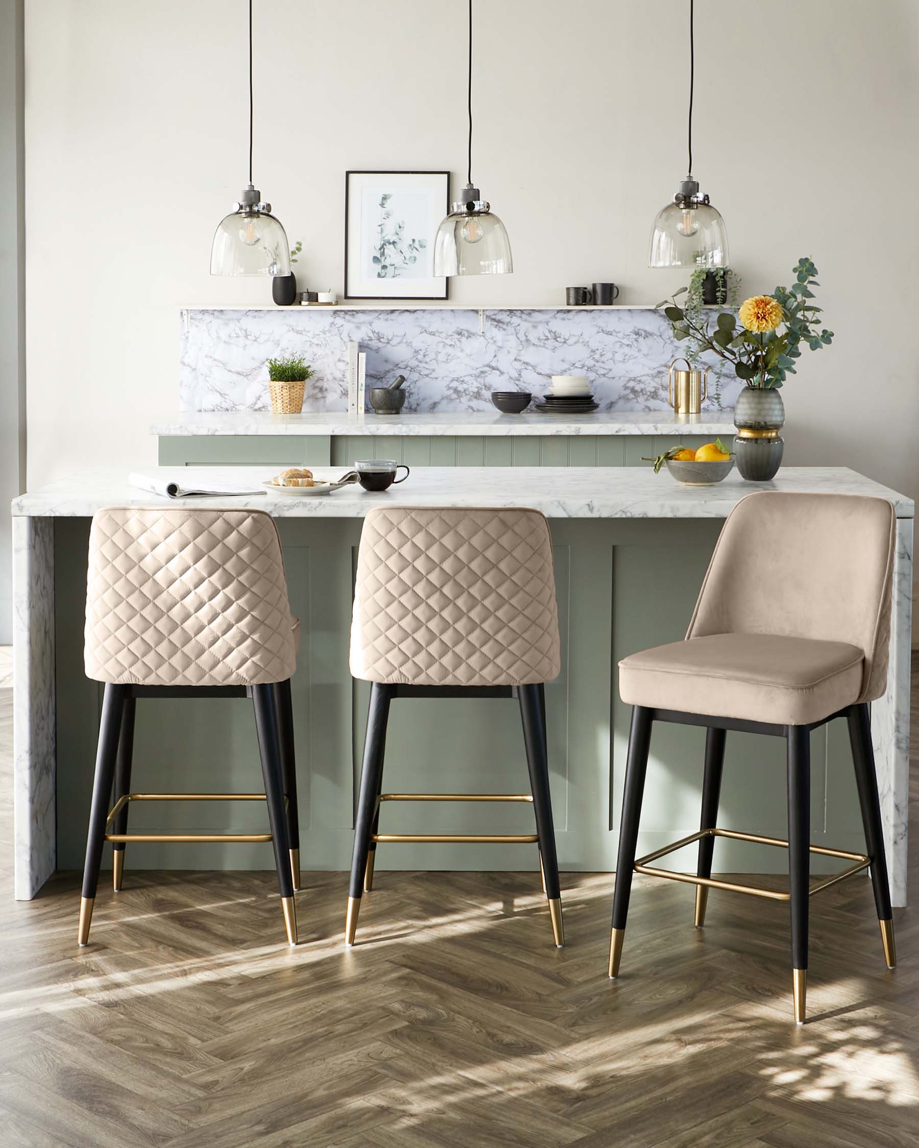 Elegant modern kitchen with a set of three stylish bar stools featuring beige diamond-tufted upholstery and black legs with gold footrests. The stools are arranged around a marble countertop kitchen island with tasteful pendant lighting overhead.