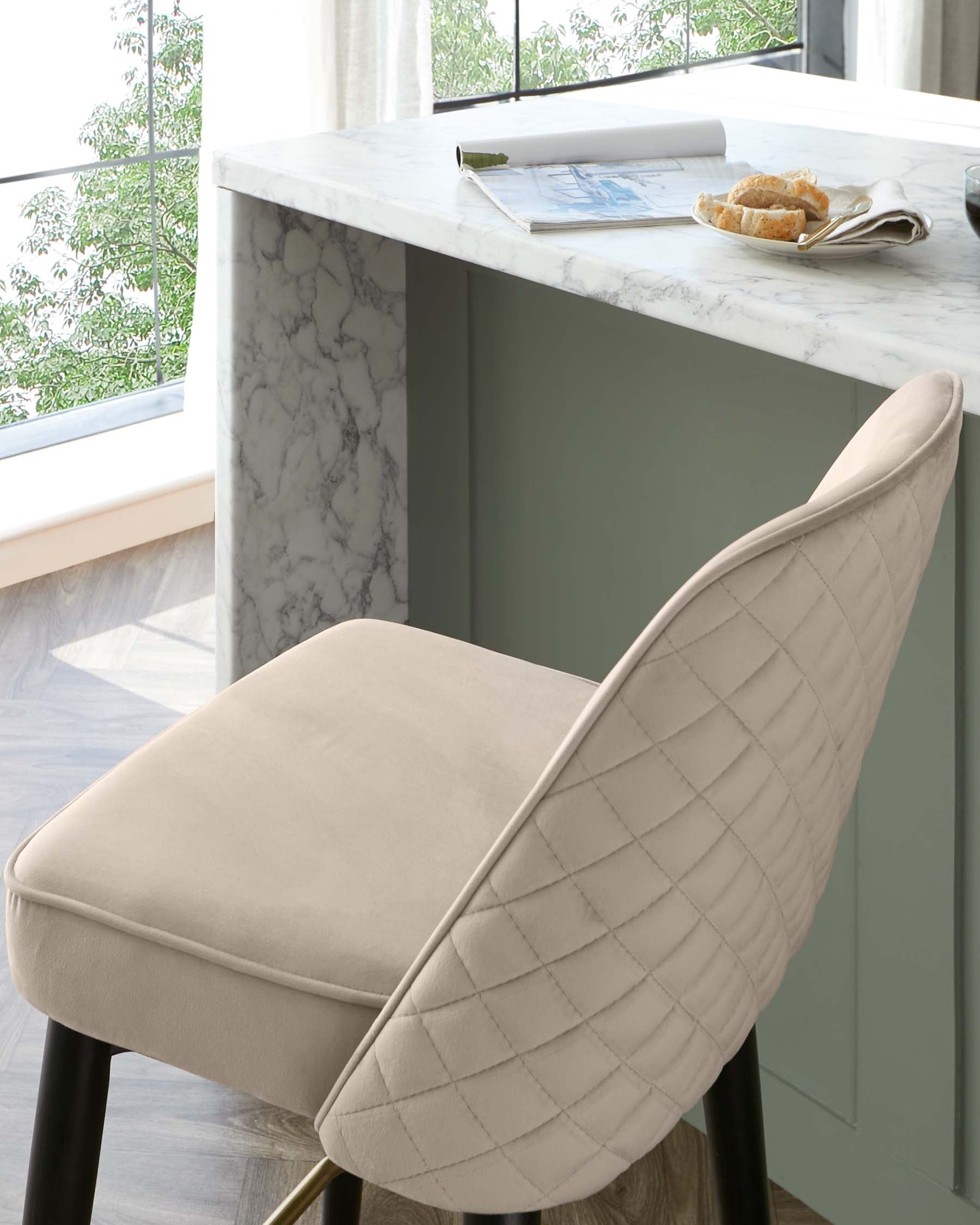 Elegant contemporary kitchen bar stool with beige upholstered seat and diamond tufted backrest, featuring sleek black legs with metallic gold footrests, juxtaposed against a white marble countertop.