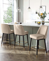 Three modern bar stools with beige diamond-tufted upholstery and black legs accented with gold footrests, positioned at a marble kitchen island under elegant pendant lights.