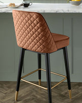 Elegant modern bar stool with quilted camel-brown upholstery and matte black legs with gold accents.