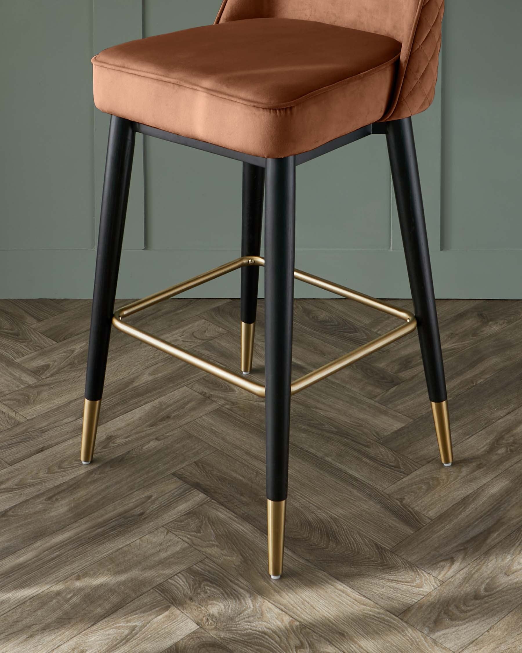 Elegant modern bar stool with a plush terracotta velvet seat and quilted backrest, mounted on sleek black tapered legs with metallic gold accents and footrest.