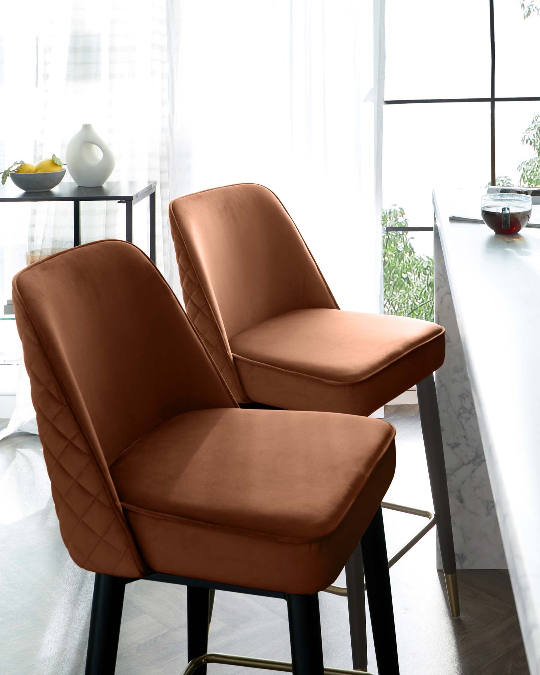 Two modern caramel brown upholstered dining chairs with quilted backrest detail and black metal legs, set in a bright, airy room with a white marble table and sheer curtains.