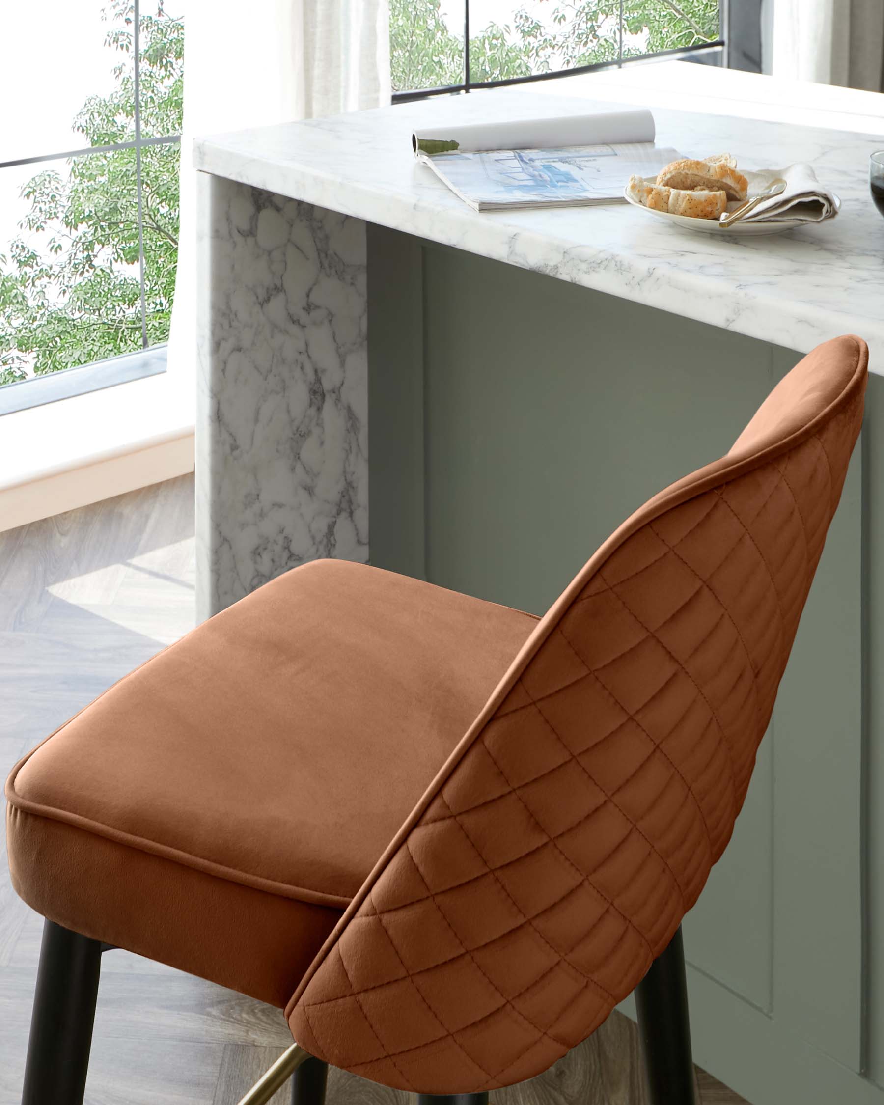 Elegant modern dining chair with burnt orange velvet upholstery and a distinctive diamond quilting on the backrest. The chair has sleek black metal legs with gold accents at the feet, paired with a marble-top kitchen island in the background.