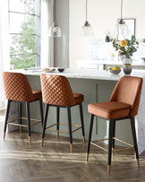 Three contemporary bar stools with caramel brown upholstered seats featuring quilted backrests, mounted on sleek black frames with brass-finished footrests, in a modern kitchen with a marble countertop.