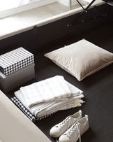 Contemporary-style black storage bed frame with a lift-up mattress revealing a light wood-coloured interior storage compartment. A patterned storage box, a beige pillow, folded textiles, and casual shoes are arranged around the bed on a dark floor, suggesting an organized and modern living space.