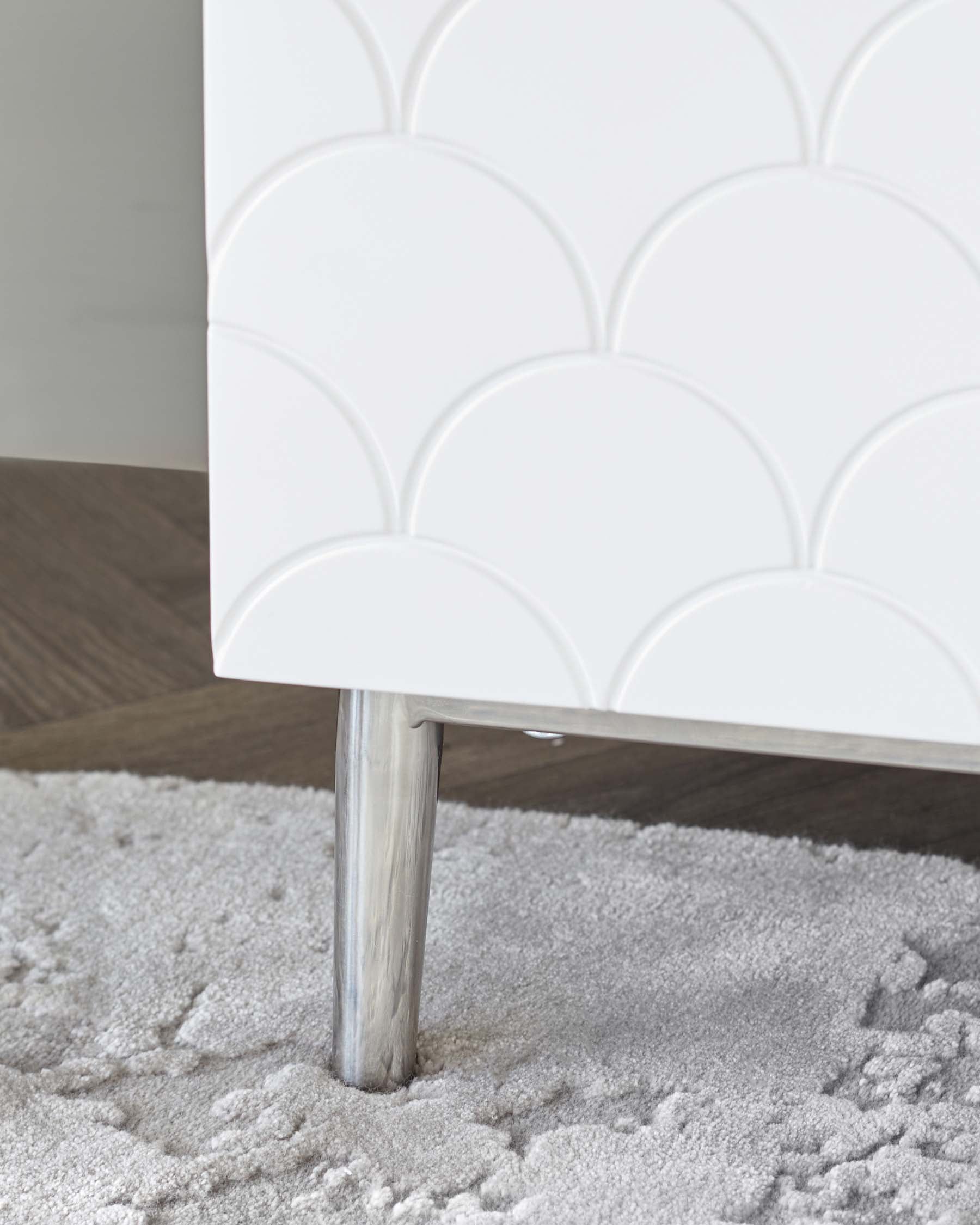 Modern white textured cabinet with scalloped pattern detail and sleek metal leg on a plush grey rug.