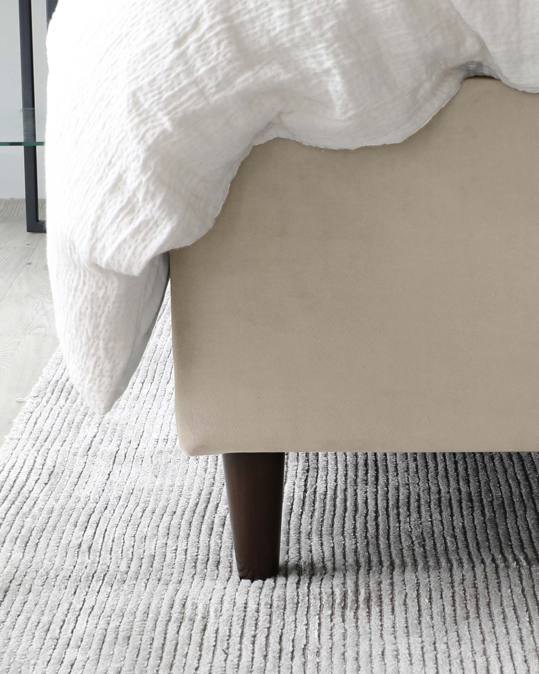 Beige upholstered bed with a white textured blanket, set against a modern room with a grey ribbed area rug and dark wooden bed leg visible, near a glass panelled mirror or window.