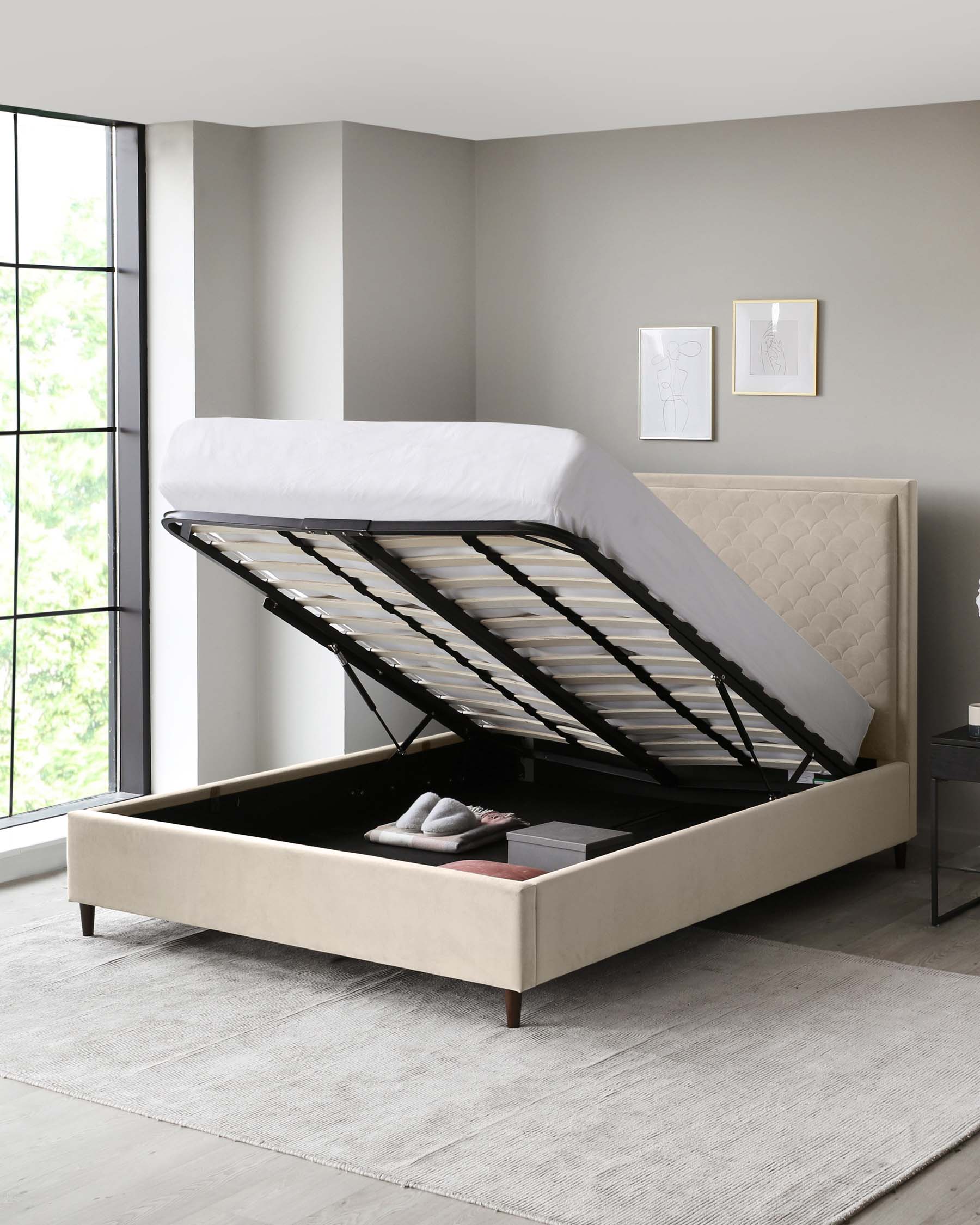 A modern beige upholstered storage bed with a lifted mattress platform revealing an under-bed storage compartment. The bed features a stylish headboard with tufting detail and is positioned on a neutral-toned area rug. A simple black bedside table is visible to the right. The scene is complemented by minimalistic artwork on the wall and natural light from a large window.