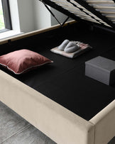 Contemporary beige upholstered storage bed with a lifted slatted base revealing a spacious black interior storage compartment, accompanied by plush decorative pillows and a warm throw blanket.