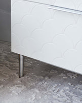 A modern white cabinet with a textured, scalloped facade and a sleek horizontal drawer handle set atop a cylindrical, brushed metal leg; showcased on a patterned grey area rug.