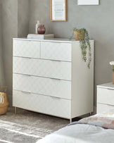 Modern white dresser with four drawers featuring a scalloped pattern design and slender metallic legs, paired with a matching two-drawer nightstand. A decorative vase, books, and a framed art piece adorn the dresser, complemented by a trailing potted plant on top. The furniture is set against a neutral grey wall on a textured area rug, creating an elegant and contemporary bedroom scene.