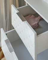 A contemporary white dresser with a scalloped pattern on the drawer fronts and sleek silver handles, partially open to reveal a light pink garment.