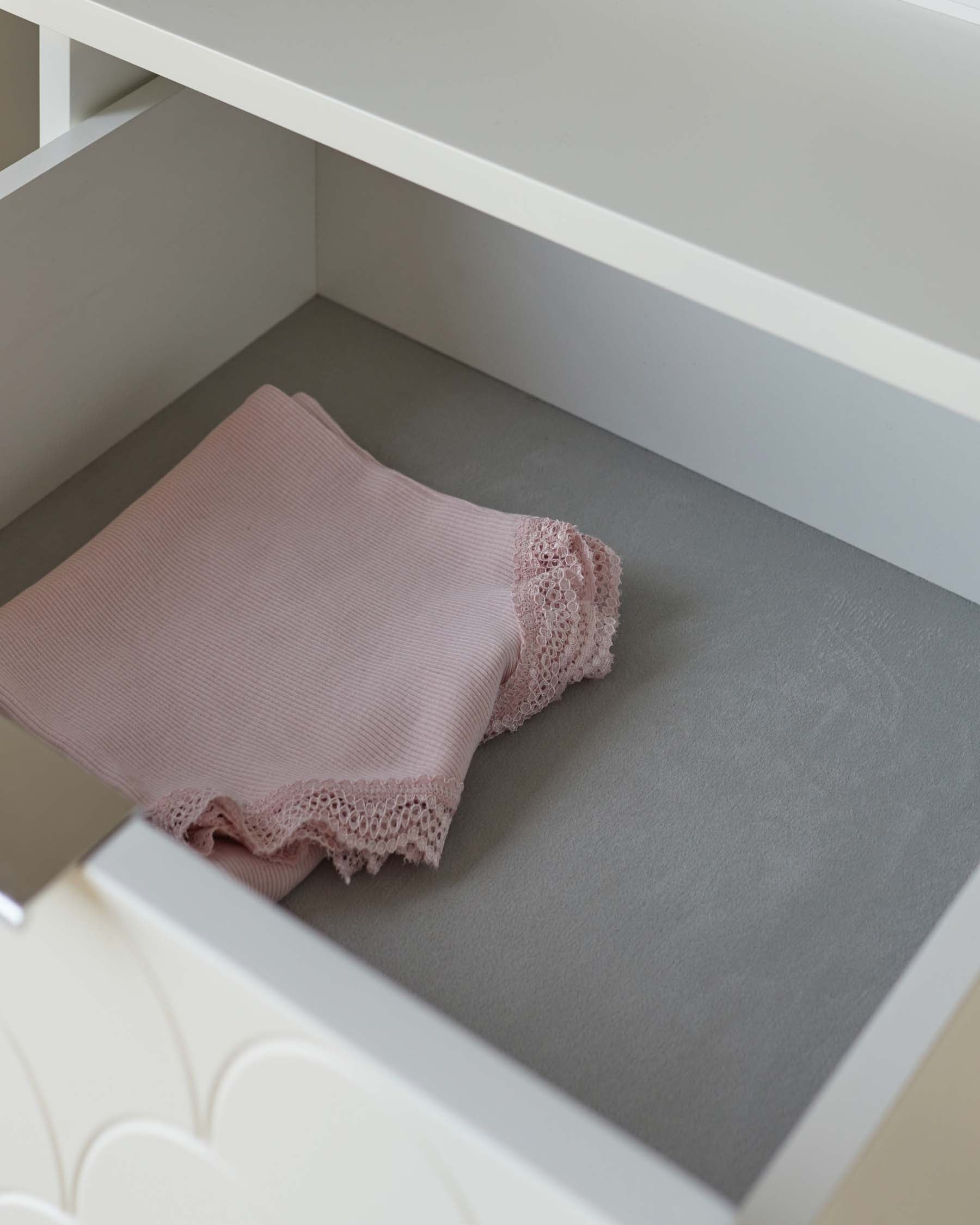 A minimalist white open drawer with a folded pink towel with lace edging lying inside. The background is a soft grey, complementing the simple yet elegant design of the furniture.