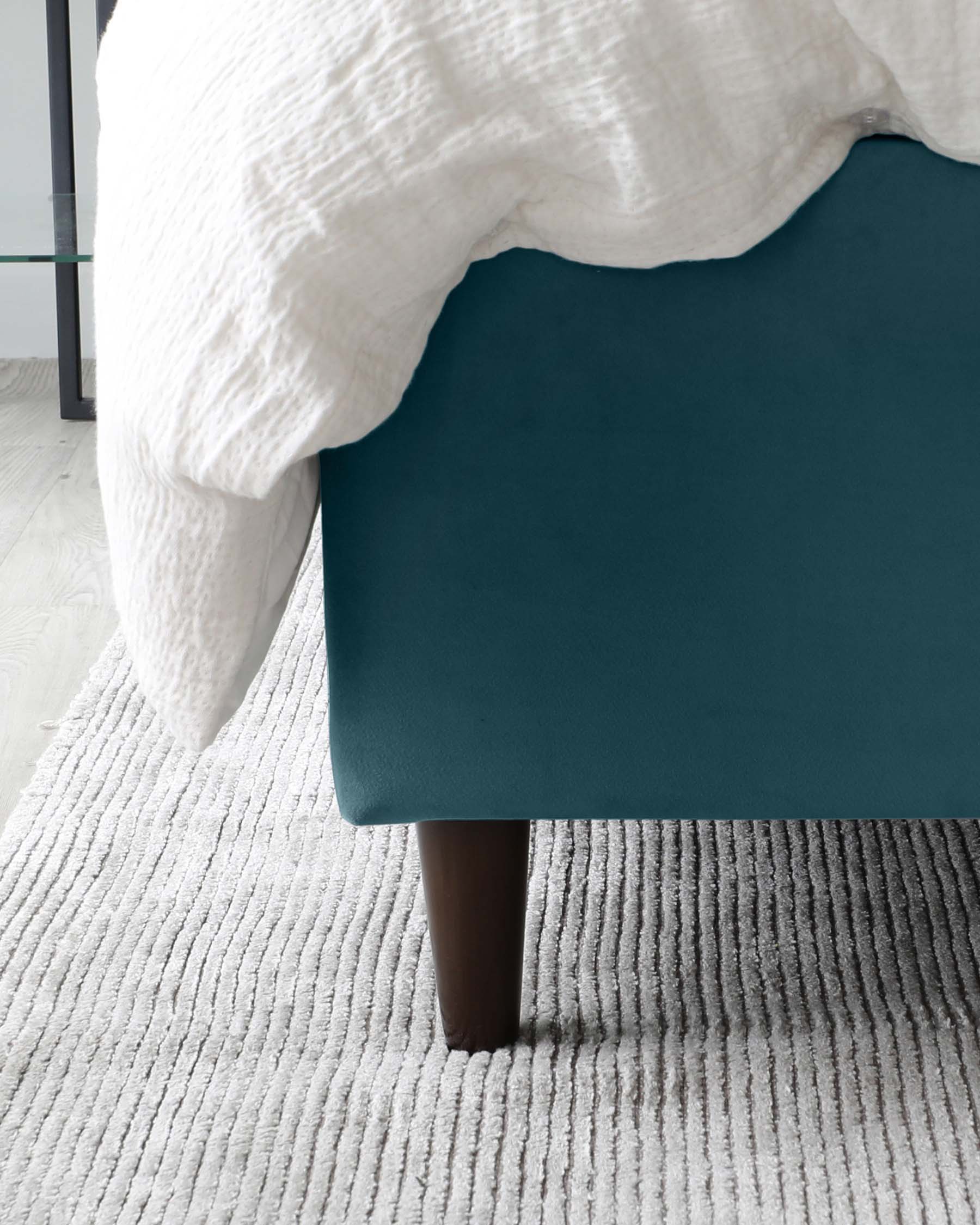 A contemporary teal upholstered bed with dark wooden legs partially covered with a white textured comforter, situated on a textured grey area rug in a light-toned room.