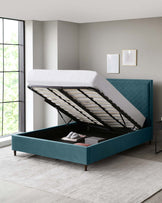 Elegant teal upholstered storage bed with a raised mattress revealing an open storage compartment, featuring a tufted headboard and dark wooden legs, set against a contemporary grey bedroom interior with minimalistic decor.