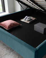 Elegant teal upholstered storage bed with the mattress platform lifted to show the spacious storage compartment beneath; on top, a pink pillow, a folded grey throw blanket, and a grey storage box accent the black mattress cover.