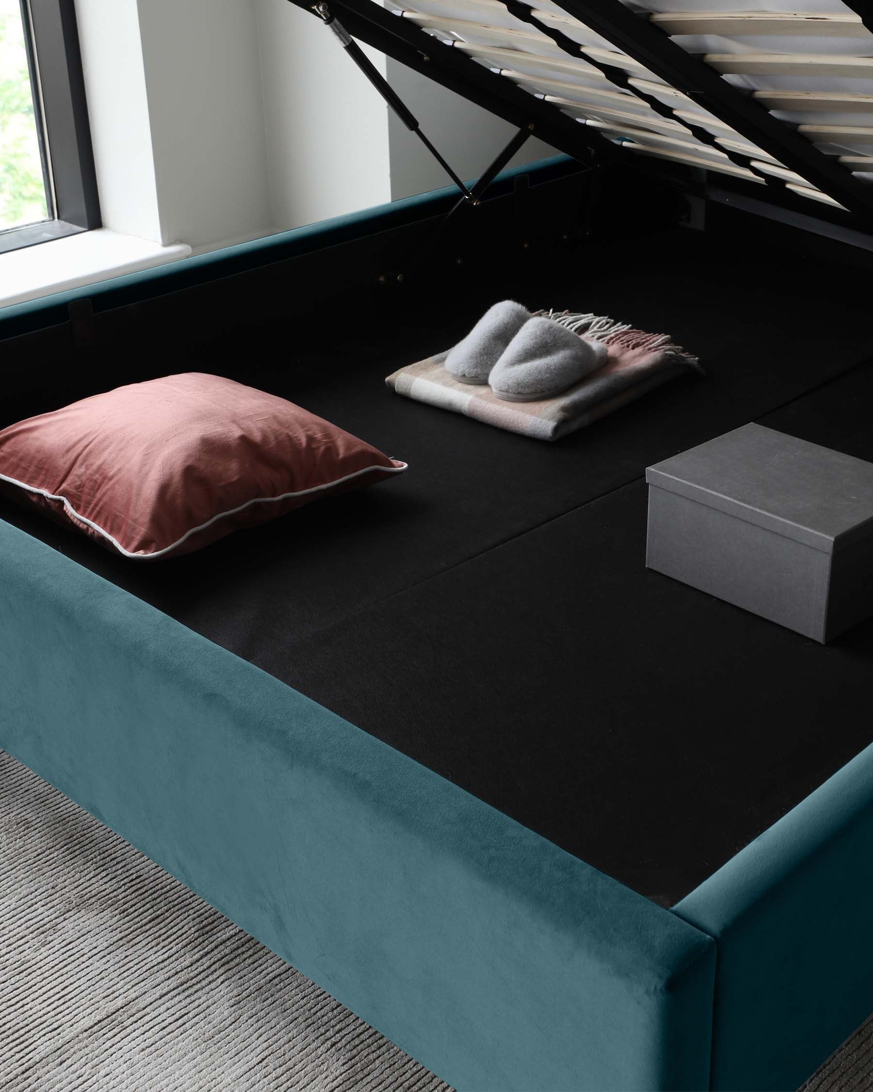 Elegant teal upholstered storage bed with the mattress platform lifted to show the spacious storage compartment beneath; on top, a pink pillow, a folded grey throw blanket, and a grey storage box accent the black mattress cover.