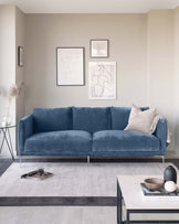 A contemporary living room featuring a plush blue three-seater fabric sofa with simple, clean lines and metallic legs. In front of it is a modern white marble-top coffee table with a black metal frame, accompanied by a sleek, black three-legged side table with a white tabletop lamp. Both tables stand over a muted, grey area rug that contrasts lightly with the pale wooden flooring.