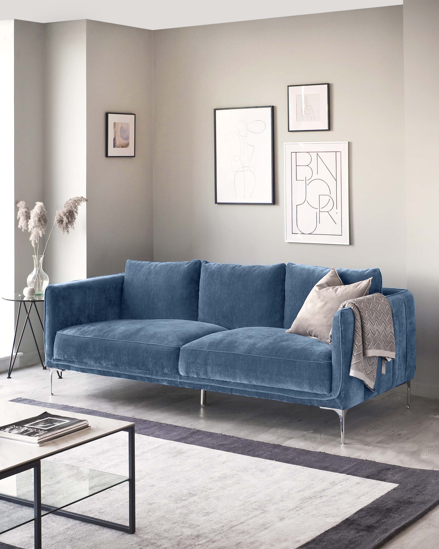 Elegant contemporary living room featuring a plush blue velvet three-seater sofa with clean lines and metal legs, a sleek glass coffee table with a metal frame, and a round side table with a black top and thin legs. The room is accented with a neutral-toned area rug and minimalist artwork on the walls.