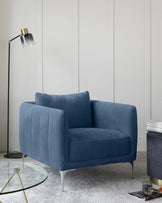 Elegant blue velvet armchair with sleek silver metal legs, featuring a plush seat, cushioned backrest, and vertical channel tufting. A contemporary floor lamp with a black stand and gold accents beside it, and a small round glass side table with a gold base in the background further complement the modern aesthetic.