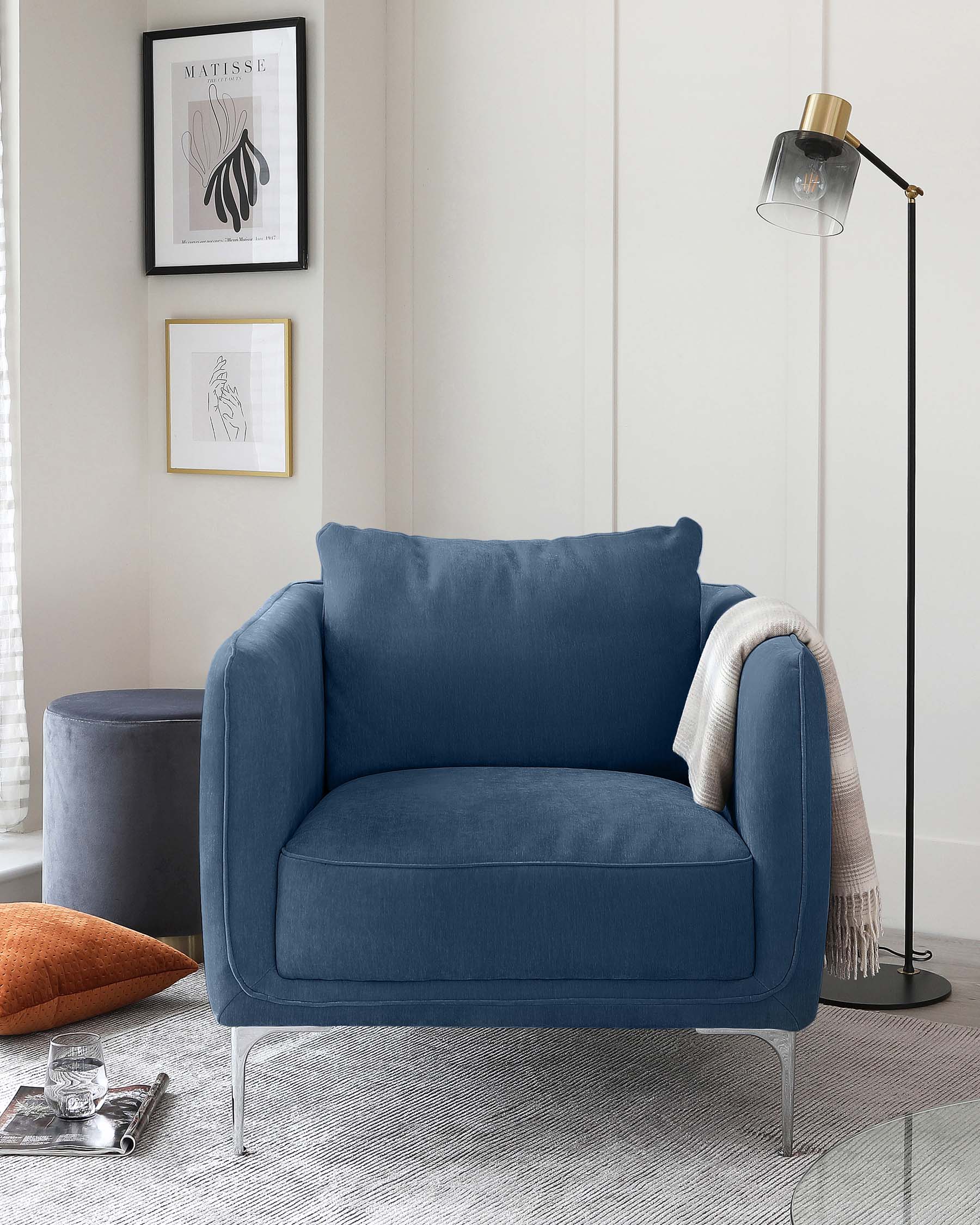 A cosy, contemporary corner with a plush, deep blue fabric armchair adorned with a soft beige throw blanket on one side. Next to the armchair is a matching blue fabric ottoman, providing a comfortable footrest or additional seating option. The ensemble is complemented by an elegant black floor lamp with a minimalist design and a transparent shade, enhancing the modern aesthetic. The setup is arranged on a textured grey rug, creating a warm, inviting space.