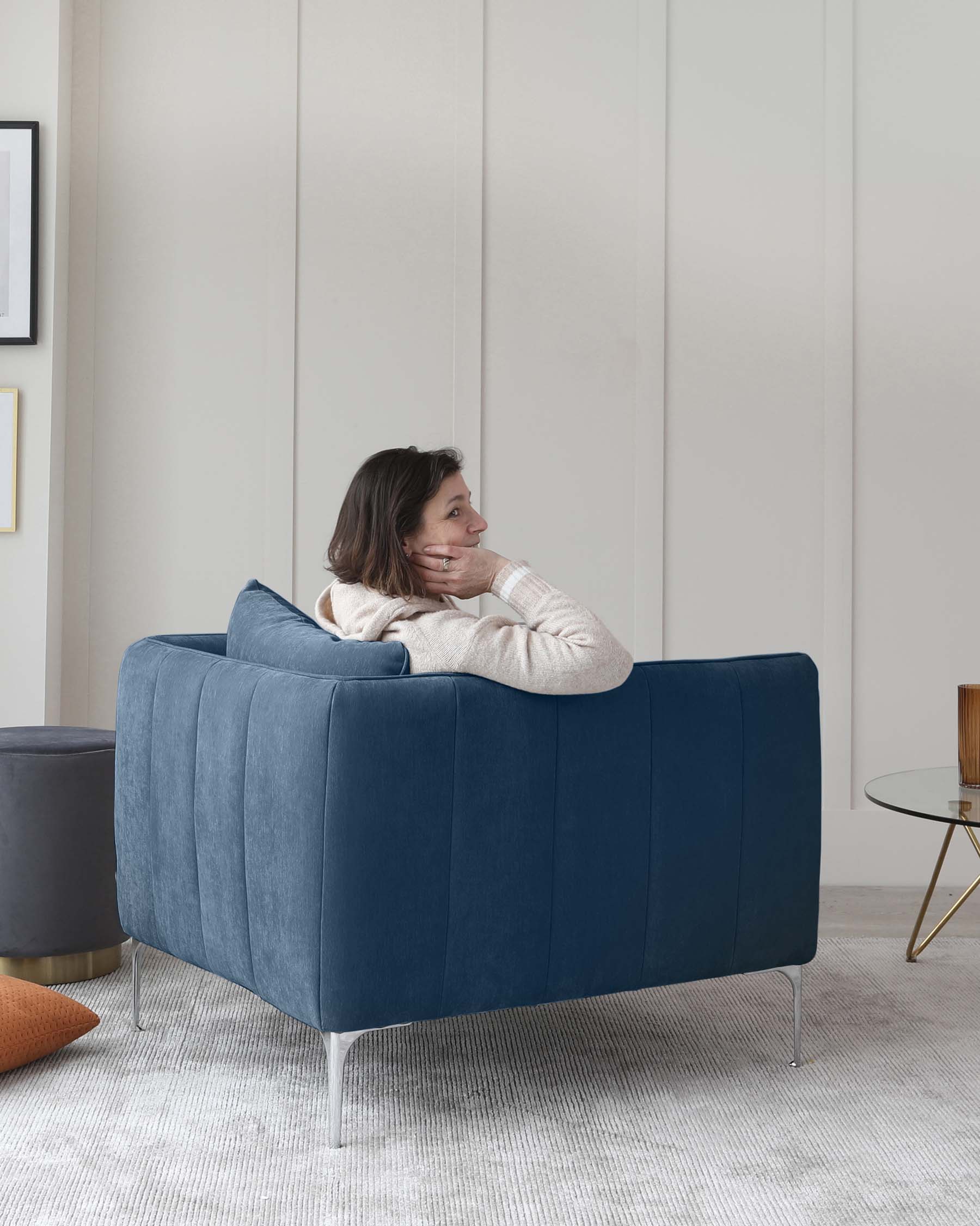 Modern velvet-upholstered loveseat in a deep blue with clean lines, rectangular armrests, and sleek metallic legs. Next to it, a round, dark-grey ottoman paired with a small side table featuring a round wooden top and thin, angular, gold-coloured metal legs.