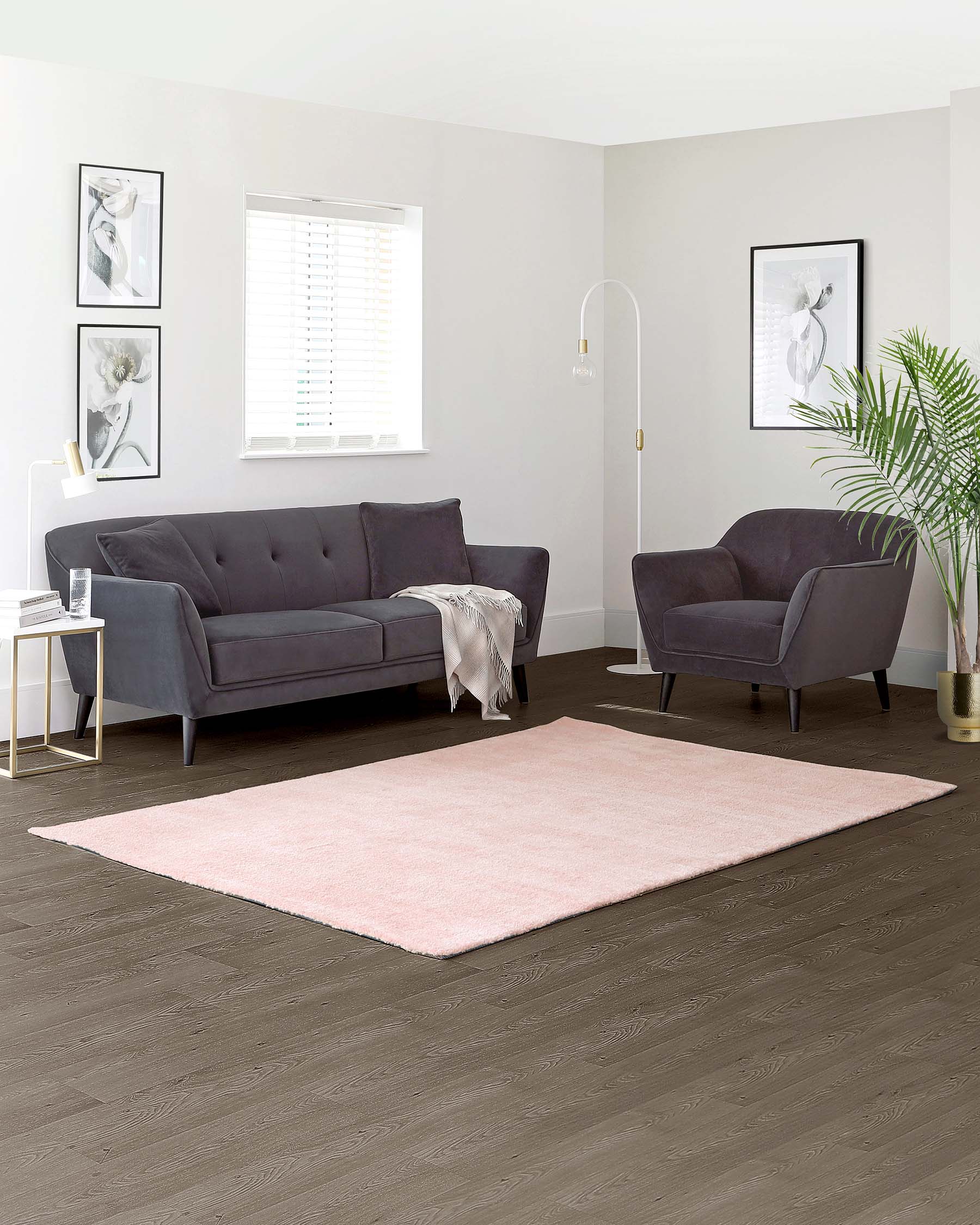 A modern living room set featuring a charcoal grey fabric sofa with tufted backrest, a matching armchair, and a white minimalist side table with gold accents. A blush pink area rug anchors the arrangement on a dark hardwood floor.