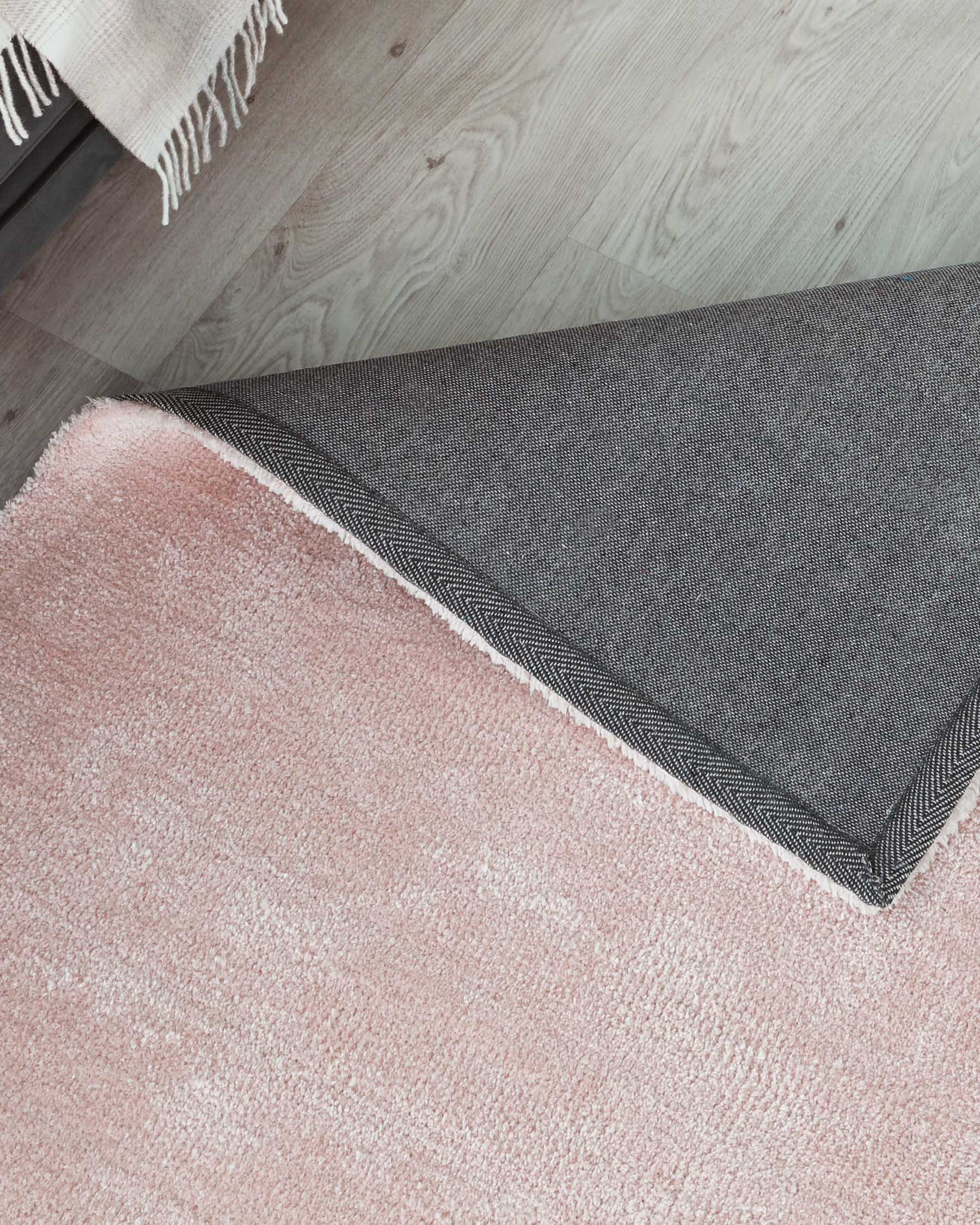 A layered rug arrangement featuring a blush pink plush rug topped with a smaller dark grey woven rug with a herringbone pattern and white fringe detail, both on a light wooden floor.