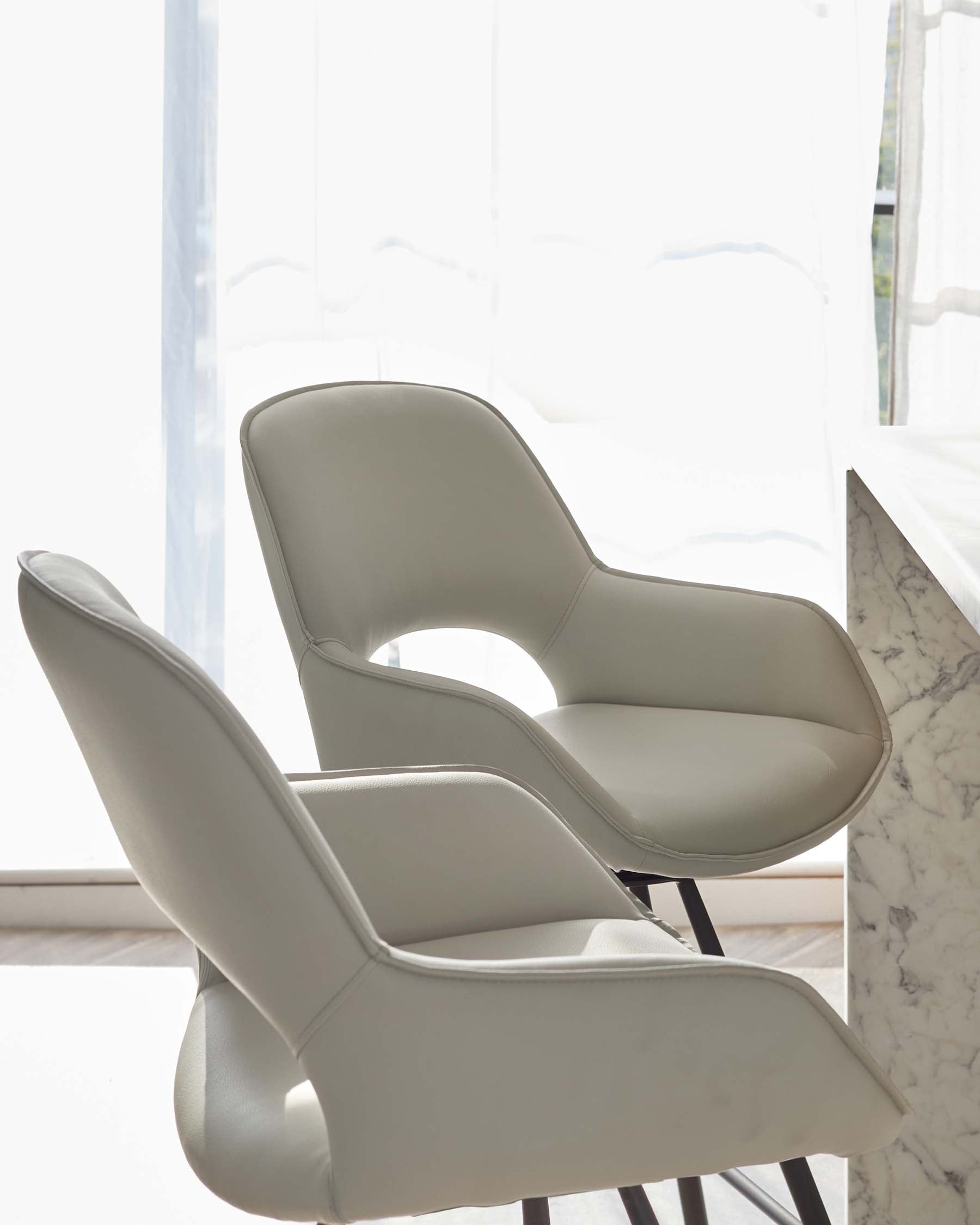 Modern light grey upholstered chairs with a smooth, curved design and dark metal legs, set against a white marble table, by a sunlit window with sheer curtains.