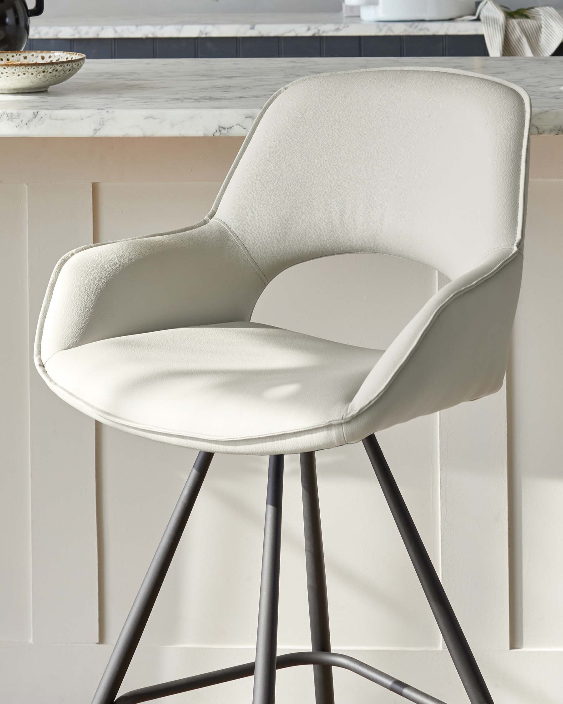 Modern beige upholstered armchair with a rounded backrest and a metal base with four angled legs, displayed in an airy interior with a marble countertop in the background.