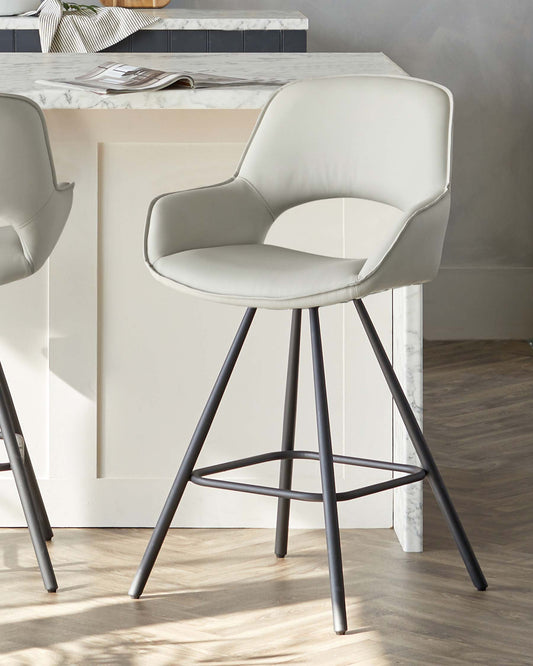 Modern light grey upholstered bar stool with a sweeping curved backrest and a comfortable, rounded seat pad, mounted on a sleek black metal frame with four slender legs and a circular footrest. The stool is positioned near a kitchen island with a white marble countertop.