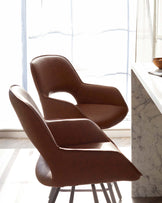 A pair of elegant brown leather mid-century modern-style chairs with a smooth finish and curved backrest. They have slim, sturdy legs that appear to be made of metal, possibly in a dark finish. The chairs are positioned next to a white marble countertop, suggesting a contemporary and stylish interior setting with natural lighting from a window with sheer curtains in the background.