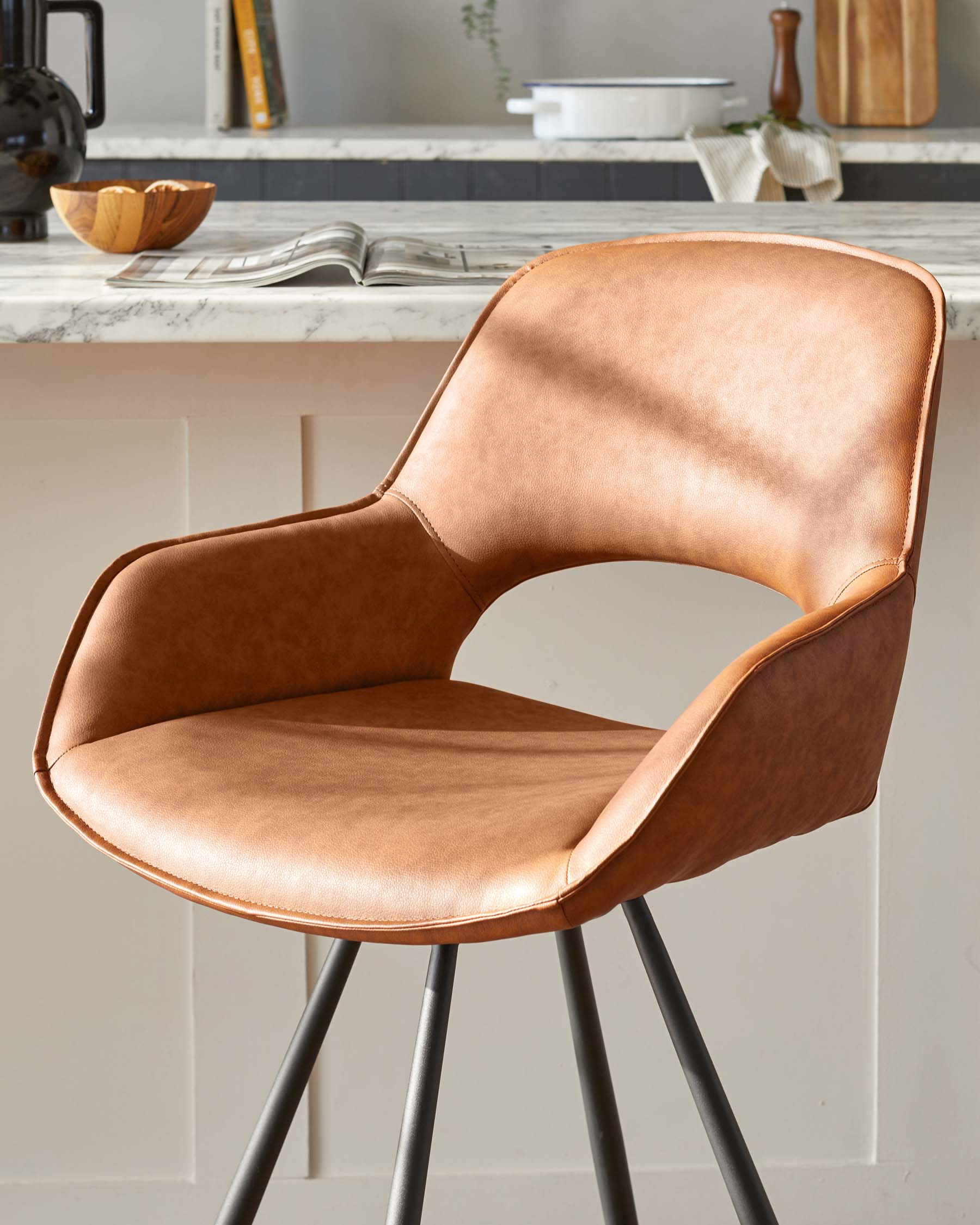 Modern caramel brown faux leather upholstered bar stool with a curved backrest and seat, featuring elegant stitching details and supported by four slender black metal legs.