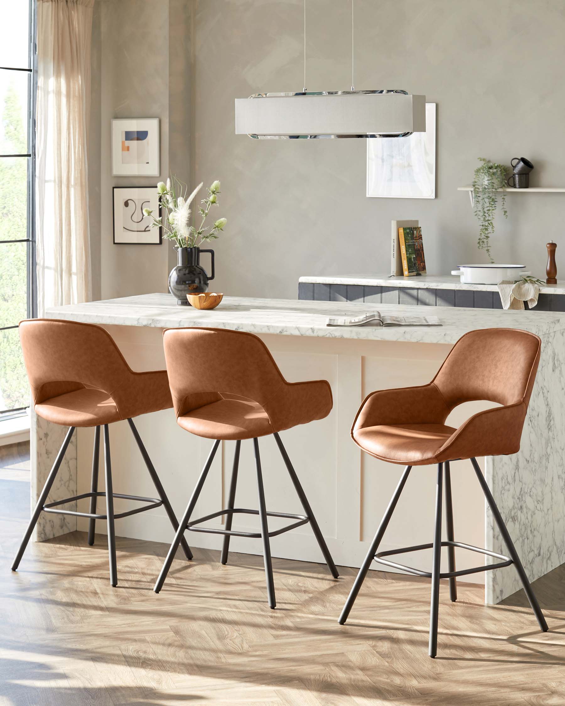 Modern kitchen interior showcasing a set of three elegant caramel-coloured upholstered bar stools with supportive backrests, armrests, and sleek black metal legs with footrests, complementing a white marble kitchen island/bar in a room with natural light.