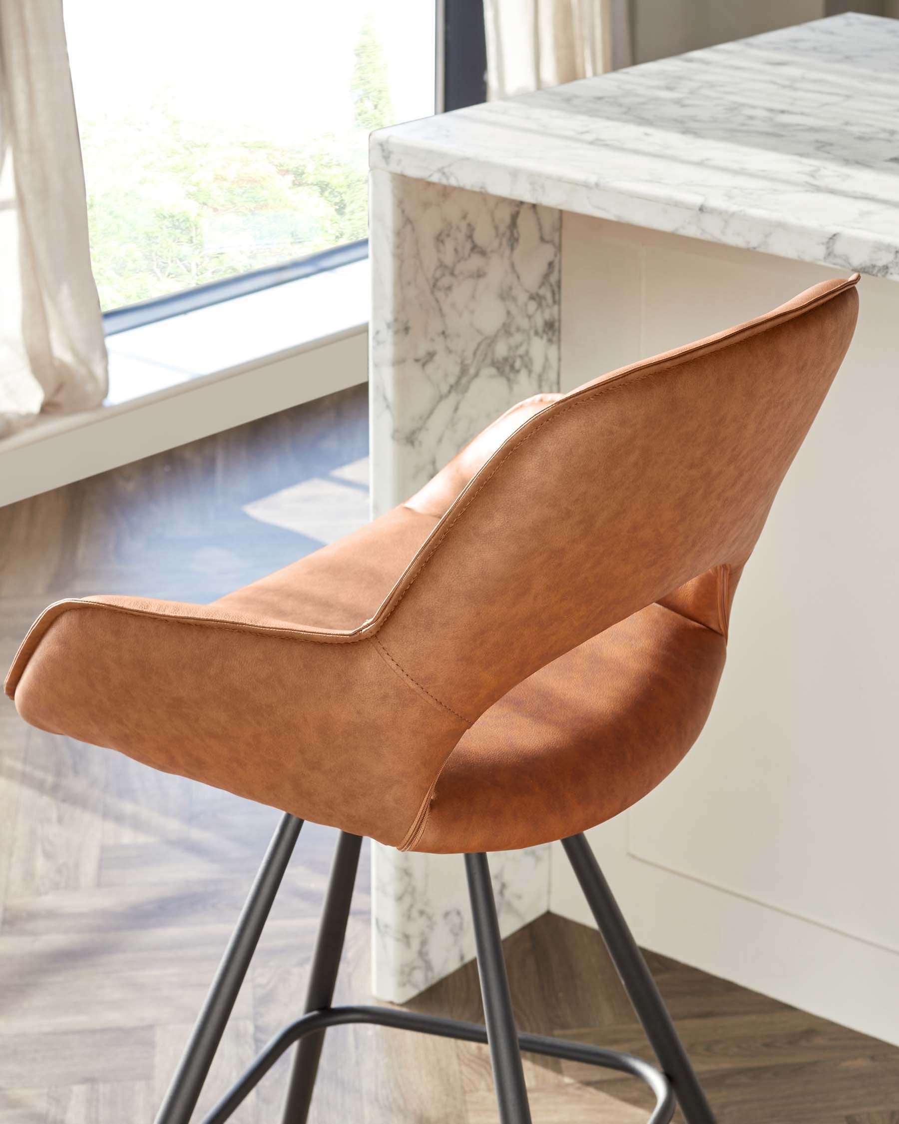 Modern, mid-height barstool with burnt orange faux leather upholstery and ergonomic design featuring a contoured back and seat. It stands on a set of sleek, black metal legs with a footrest, positioned by a kitchen island with a white marble countertop.