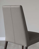 Modern dining chair with a high back design, featuring sleek, grey faux-leather upholstery and a subtle vertical stitch detail running down the centre of the backrest. The chair is supported by dark wooden legs with a slightly tapered shape, providing a contrast to the light upholstery. The image showcases the chair against a neutral background, emphasizing its clean lines and elegant silhouette.