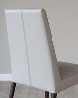 Elegant modern dining chair featuring a white leatherette upholstery with a subtle vertical stitching on the high backrest and a dark wooden frame with straight rear legs and slightly outward-angled front legs.