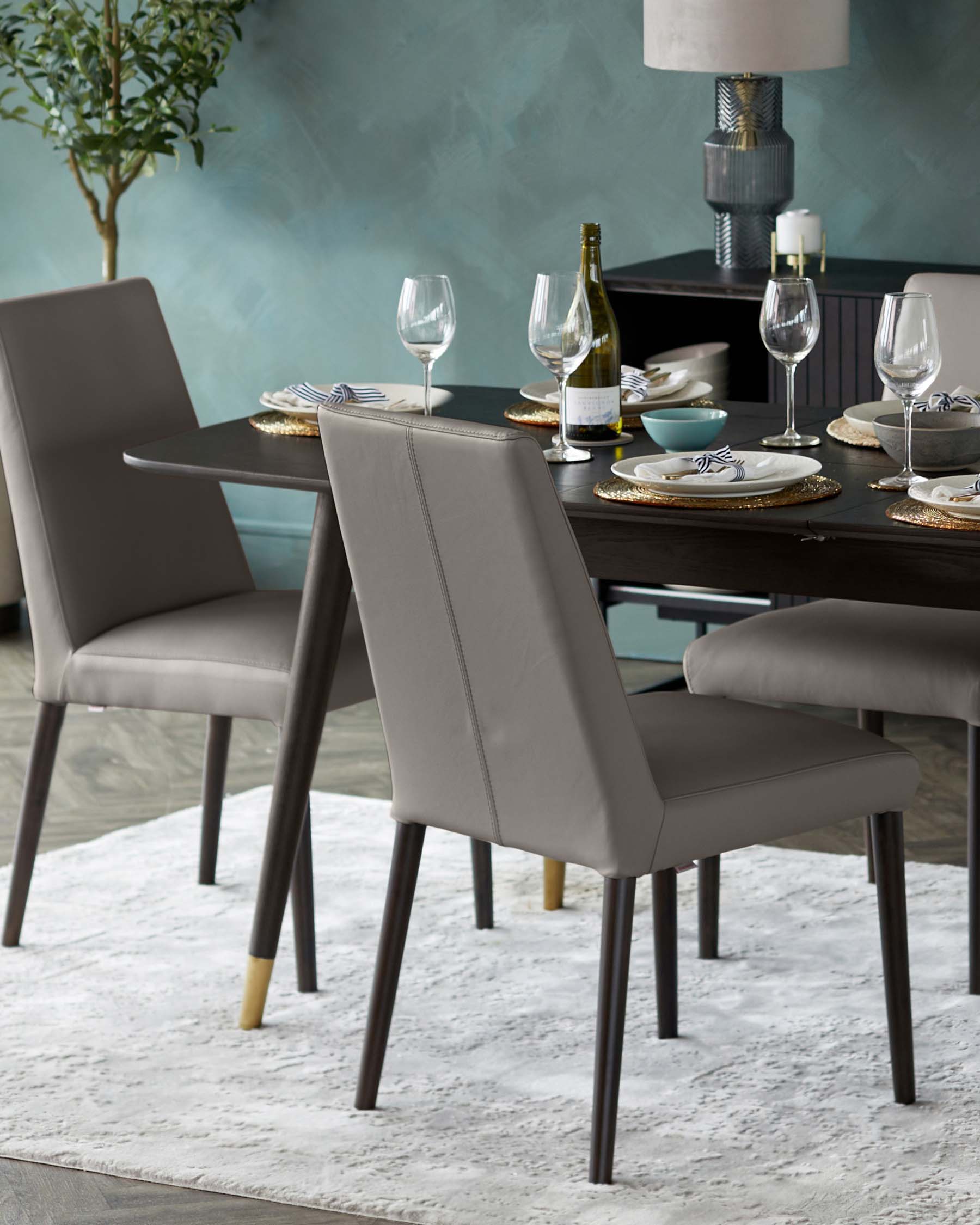 A contemporary dining set featuring an elegant dark wood table and four matching chairs with sleek grey upholstery. The chairs have a clean, minimalist design with high backs and tapered legs, some with subtle gold-coloured tips, complementing the table's straight lines and modern aesthetic. The table is set with fine dinnerware, wine glasses, and a bottle, suggesting a sophisticated dining experience. A plush area rug underneath adds a layer of texture to this stylish ensemble.