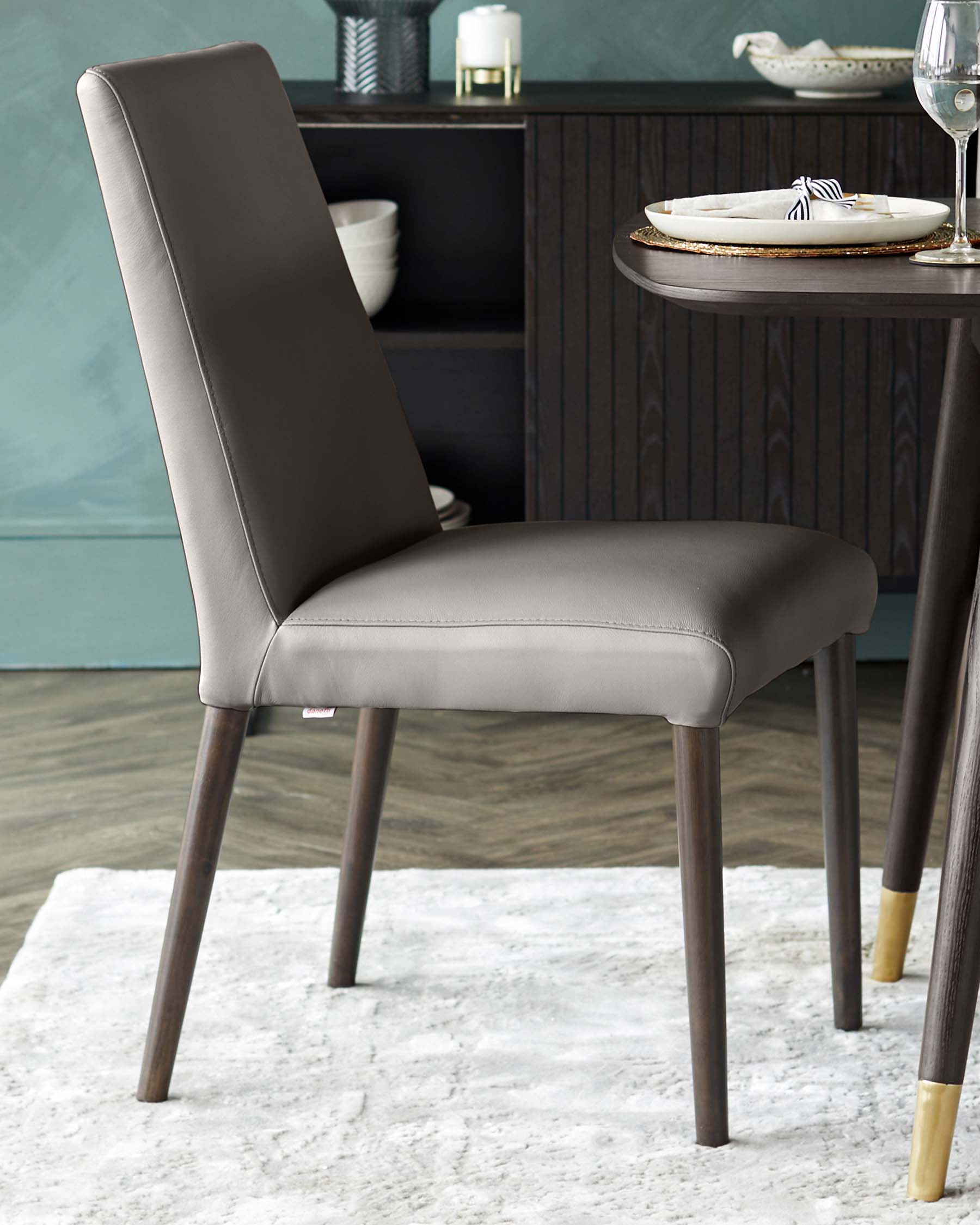 Modern dining chair with a sleek silhouette, upholstered in grey faux leather, featuring a high back and dark wooden legs with metallic gold accents at the feet.