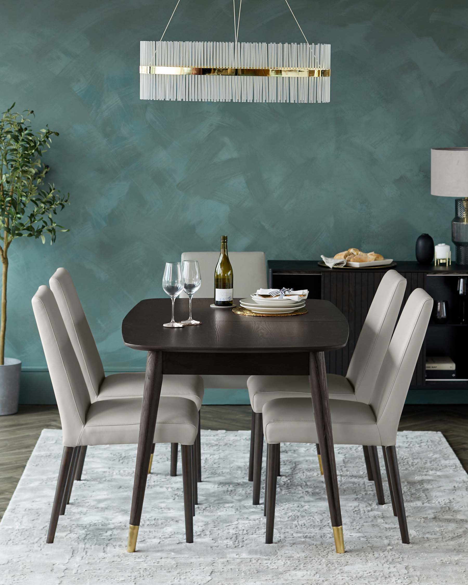 Elegant contemporary dining room set featuring a dark brown circular table with gold-accented tapered legs and four taupe upholstered chairs with contrast stitching and slender dark wood legs finished with gold tips. A sleek horizontal chandelier hangs above, and the scene is staged on a textured light grey area rug.