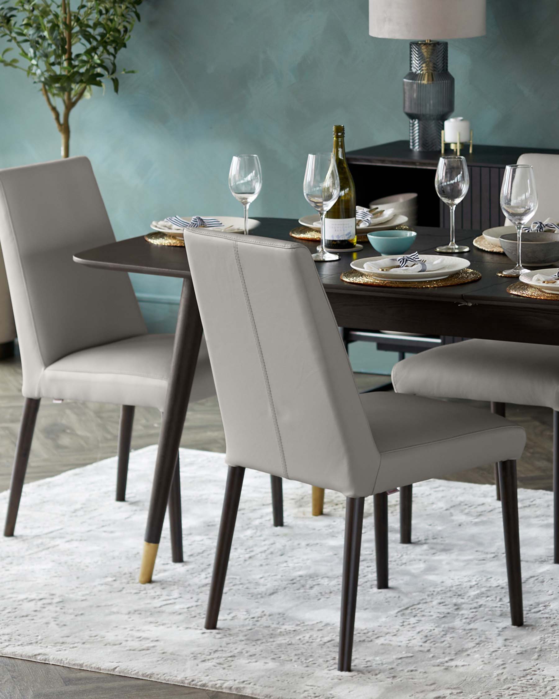 Modern dining room setting featuring a sleek, dark wood dining table complemented by elegant, light grey upholstered chairs with clean lines and contrasting dark tapered legs, some tipped with gold caps. The setting rests on a subtle white textured area rug.