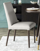 Elegant modern dining chair with a smooth, light grey upholstery and dark wooden legs, some featuring stylish gold-tone tips. A contemporary round dining table with a dark wood finish is partially visible in the background, complemented by a sophisticated dark shelving unit displaying dishware. The setting is accented by a soft teal wall and rests on a luxurious white marbled area rug.