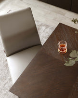 Modern dark wooden table with a textured finish, partially obscured by a taupe leather chair with a high backrest, alongside a glass of whiskey resting on the table, all set on a beige textured rug.