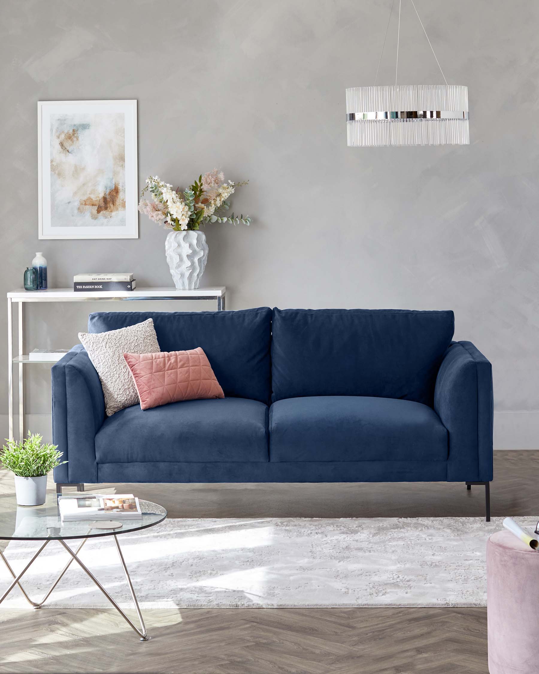 A navy blue couch with cushions, a glass coffee table, a light fixture above, and a decorative vase on a side table.