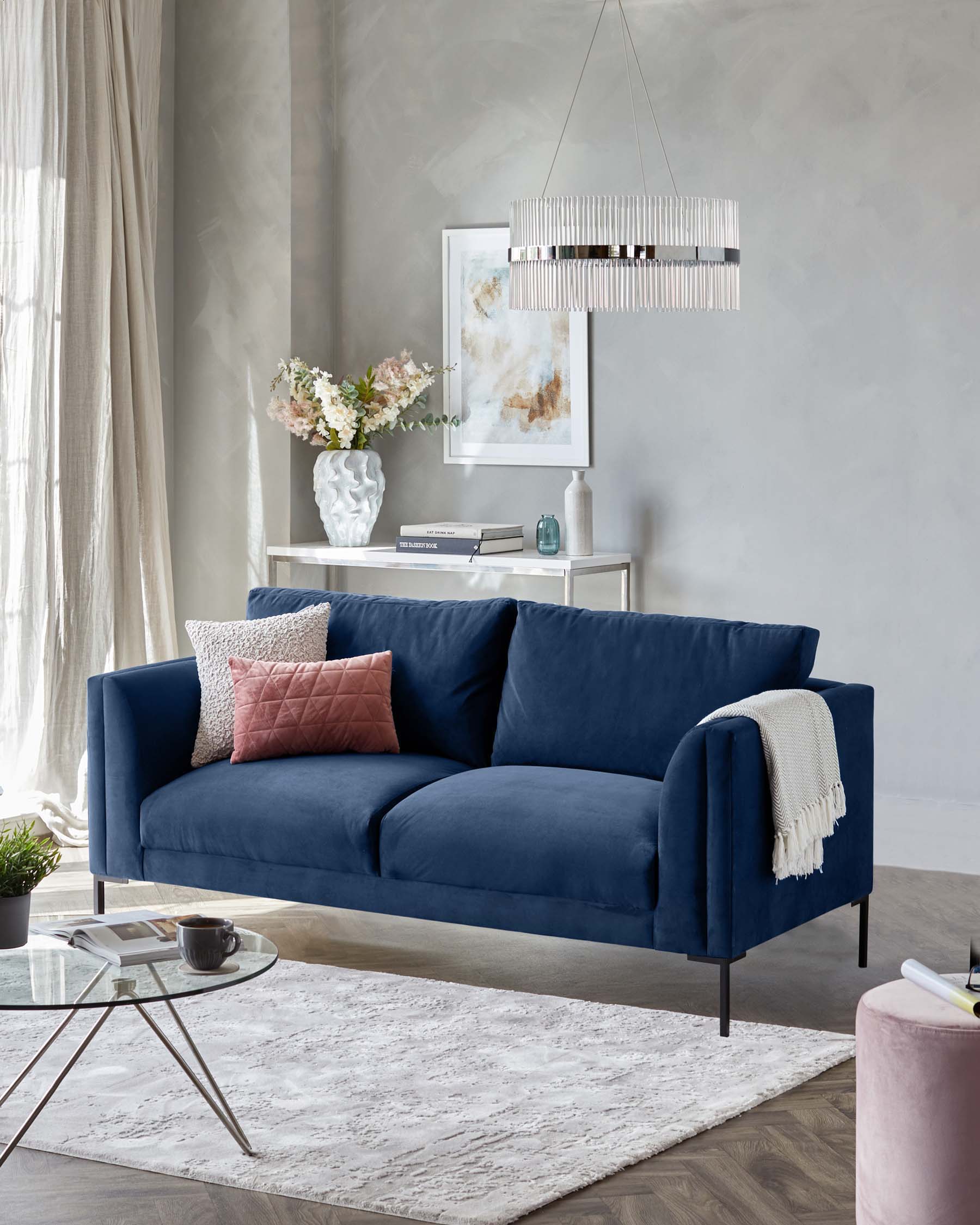 Elegant modern living room setup featuring a luxurious deep blue three-seater sofa with plush cushions, accompanied by a round glass-top coffee table with a sleek metal frame. A textured white throw blanket drapes casually over one side of the sofa, adding a cosy touch to the clean lines of the furniture. A blush pink round ottoman sits off to the side, providing additional seating or a spot to rest. The room is anchored by a soft white area rug with a subtle pattern, creating a warm and inviting ambiance.