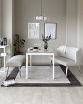 Modern minimalist furniture set featuring a white rectangular dining table with a sleek finish accompanied by a white upholstered bench and a complimentary two-seater sofa, all poised on a layered two-tone grey area rug. A slim white side table with a lower shelf, accessorized with decorative items, stands to the left, enhancing the contemporary aesthetic.